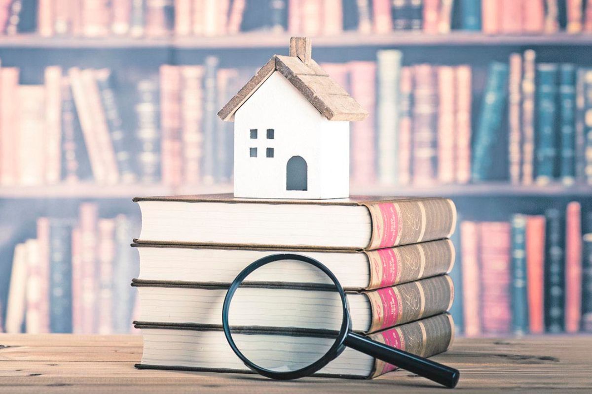 Kleines Modellhaus steht auf einem Stapel Bücher mit einer Lupe davor, im Hintergrund ein unscharfes Bücherregal.