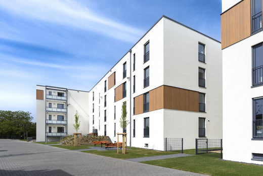 Modernes mehrstöckiges Wohngebäude mit weißer Fassade und Holzverkleidungselementen, umgeben von grünen Außenflächen und einem Spielplatz unter blauem Himmel.