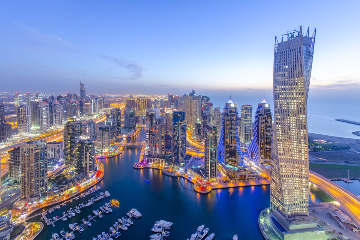 Ein beeindruckendes Panorama von Dubai Marina, beleuchtet von der nächtlichen Skyline. Die hoch aufragenden Wolkenkratzer, darunter der markante verdrehte Cayan Tower, reflektieren die Lichter der Stadt.