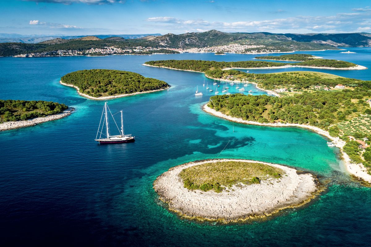 Strahlendes, türkisfarbendes Wasser zwischen mehreren kleinen Inseln im Meer. Ein Segelboot schippert auf dem Wasser.