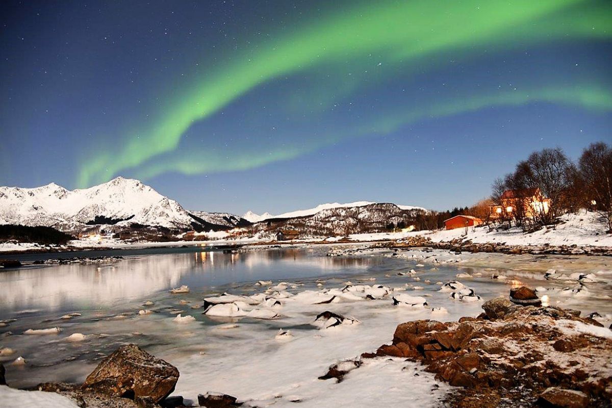 Nordlichter bilden einen grünen Bogen über einer verschneiten Winterlandschaft mit Bergen im Hintergrund. Ein kleines Dorf mit beleuchteten Häusern spiegelt sich im teilweise gefrorenen Fluss im Vordergrund.