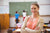 Eine Lehrerin mit lachsfarbenem T-Shirt und beigem Cardigan. Im Hintergrund Kinder, die an die Tafel mahlen und an ihrem Platz sitzen.