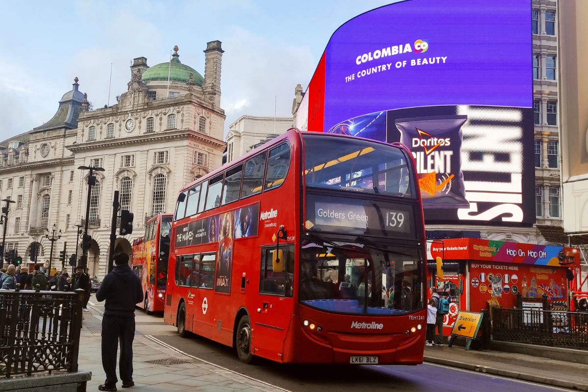 Ein typischer roter Doppeldeckerbus fährt durch London. Im Hintergrund sind die berühmten leuchtenden Werbetafeln zu sehen, Menschen bewegen sich durch den belebten Platz, umgeben von historischen Gebäuden und moderner Werbung.