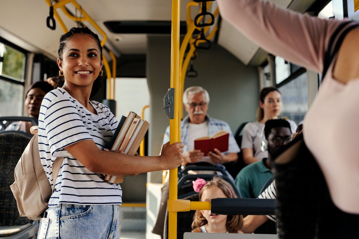 Alt-Text:  Eine junge Frau steht lächelnd in einem gut besuchten Bus, hält Bücher in der Hand, während andere Passagiere sitzen und lesen.