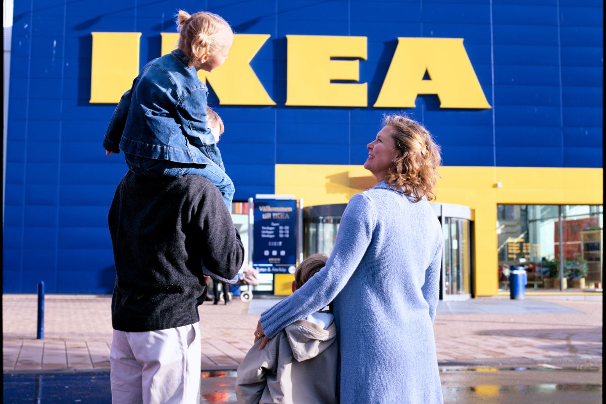Eine Familie steht vor einem IKEA-Eingang, der Mann trägt ein Kind auf den Schultern, die Frau hält ein weiteres Kind. Alle blicken auf das Geschäft.