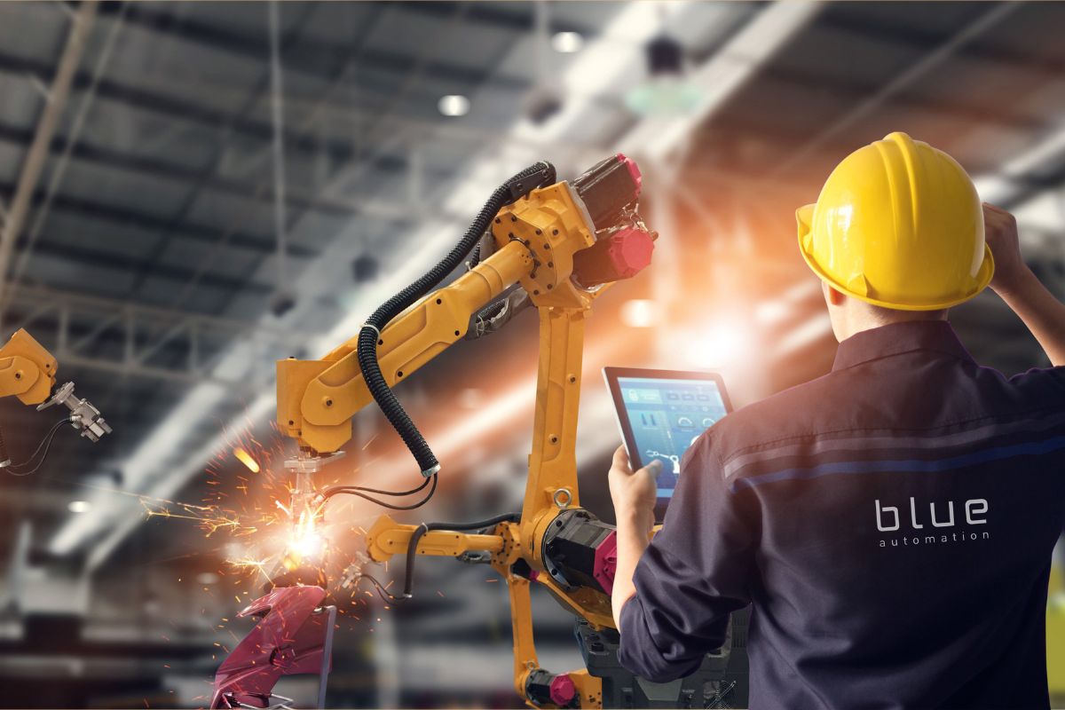 Ein Ingenieur, der in einer Fabrikhalle mit einem Tablet arbeitet, steuert gelbe Roboterarme, die Funken beim Schweißen erzeugen.