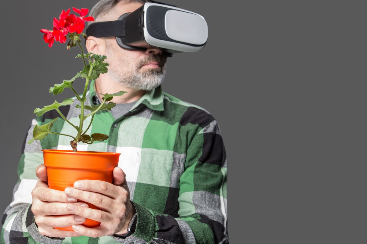 Ein Mann mit grauem Bart und grünem karierten Hemd trägt eine Virtual-Reality-Brille und hält einen orangen Blumentopf mit einer roten Geranienpflanze vor sich.