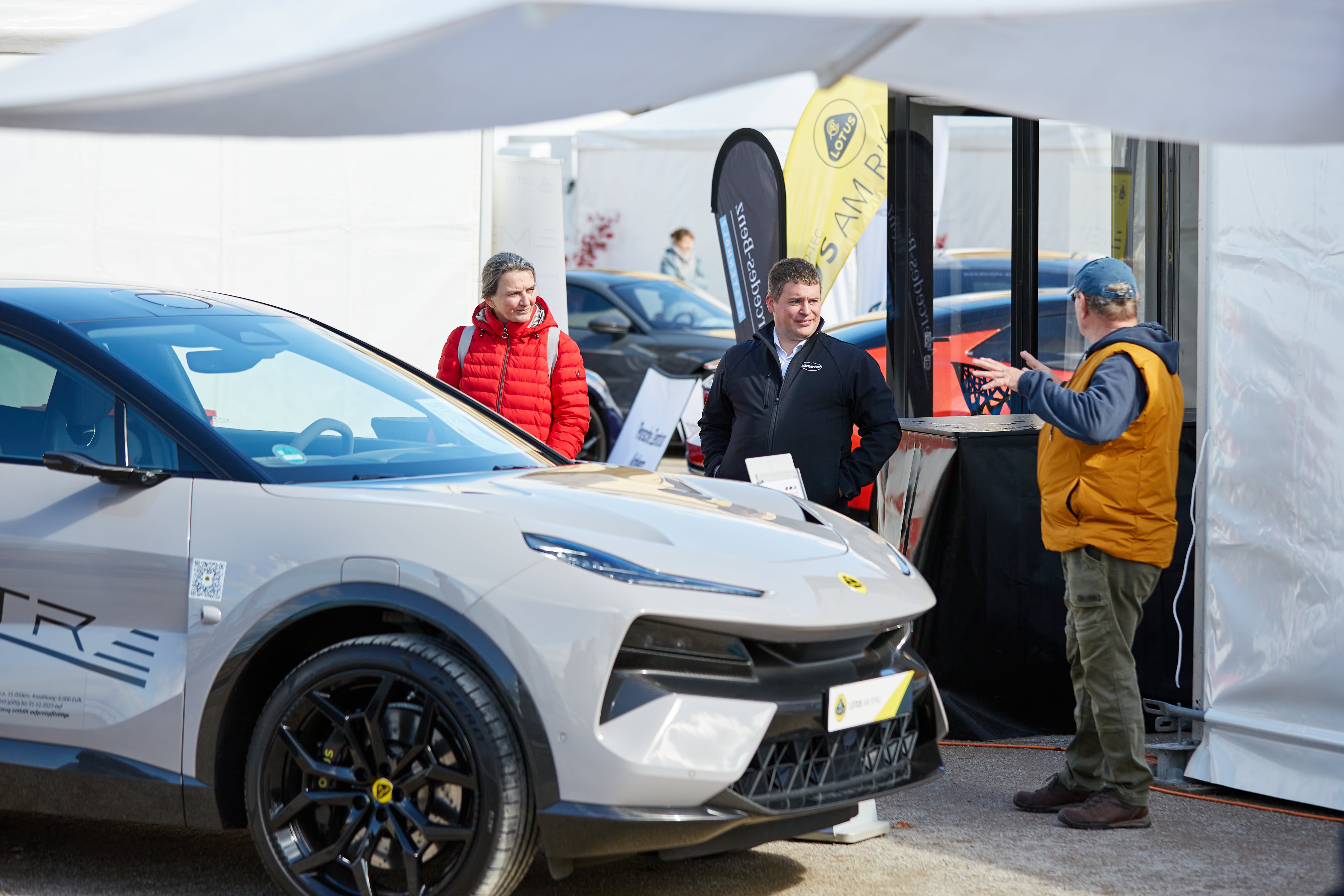 Eine Gruppe von drei Personen steht neben einem modernen, grauen Elektroauto. Eine Person in einer gelben Weste spricht gestikulierend, während die anderen beiden aufmerksam zuhören.