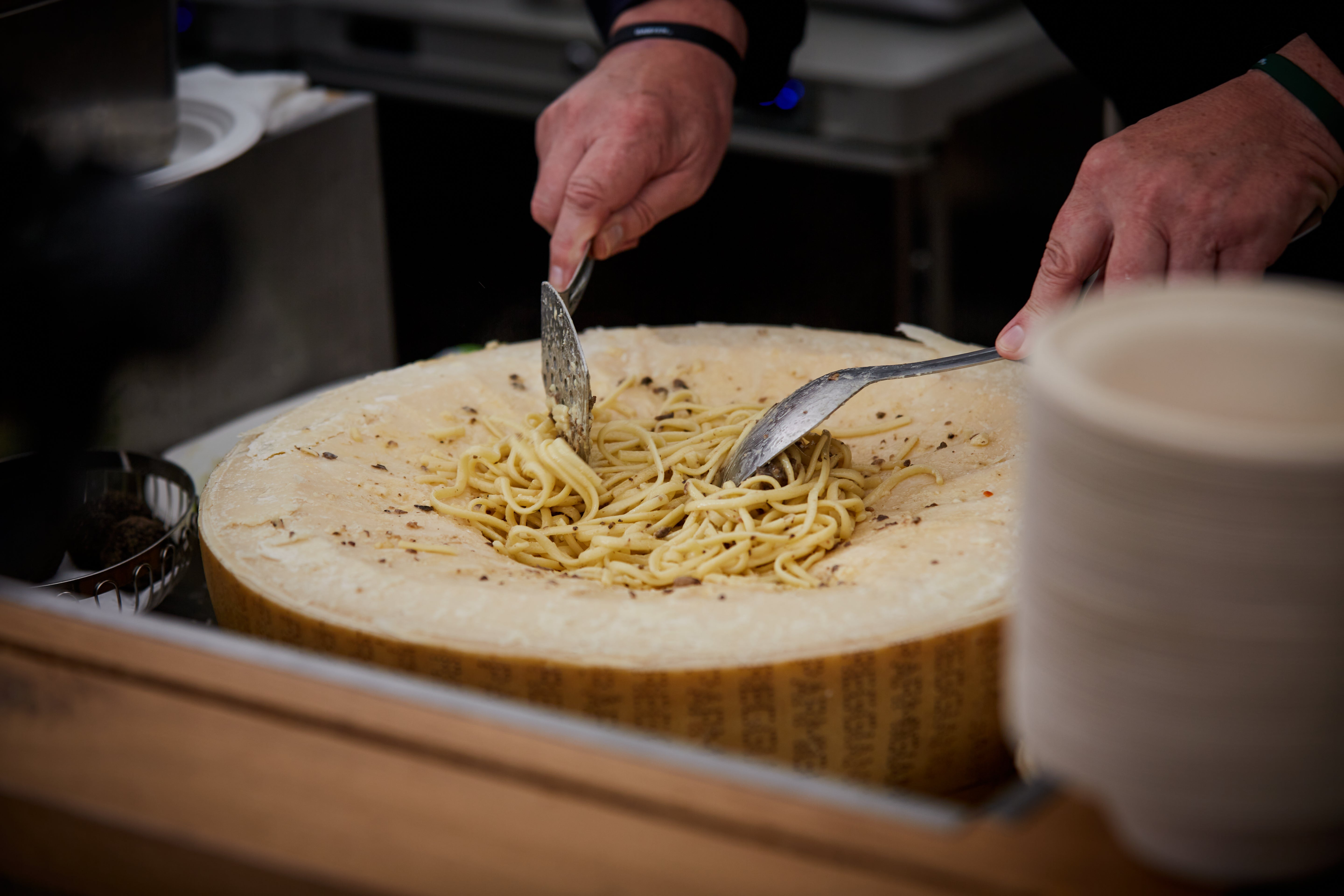 Nahaufnahme von Händen, die Spaghetti mit zwei Löffeln in einem großen Parmesanlaib mischen. Die cremige Sauce wird direkt im Käse angerührt, um den Nudeln Geschmack zu verleihen.