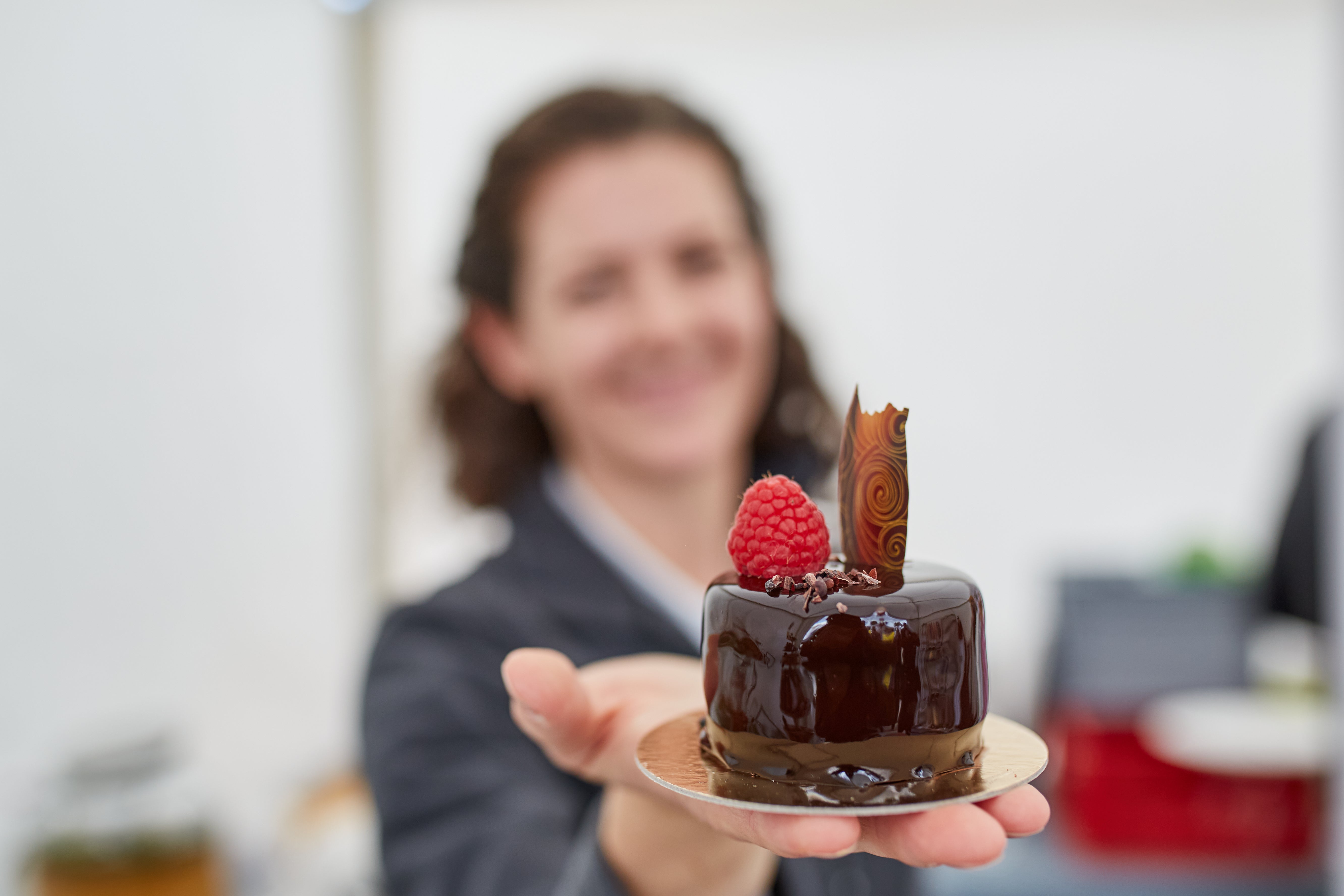 Eine Frau im Hintergrund hält eine glänzende Schokoladentorte in der Hand, dekoriert mit einer Himbeere und einem kunstvollen Schokoladenornament. 