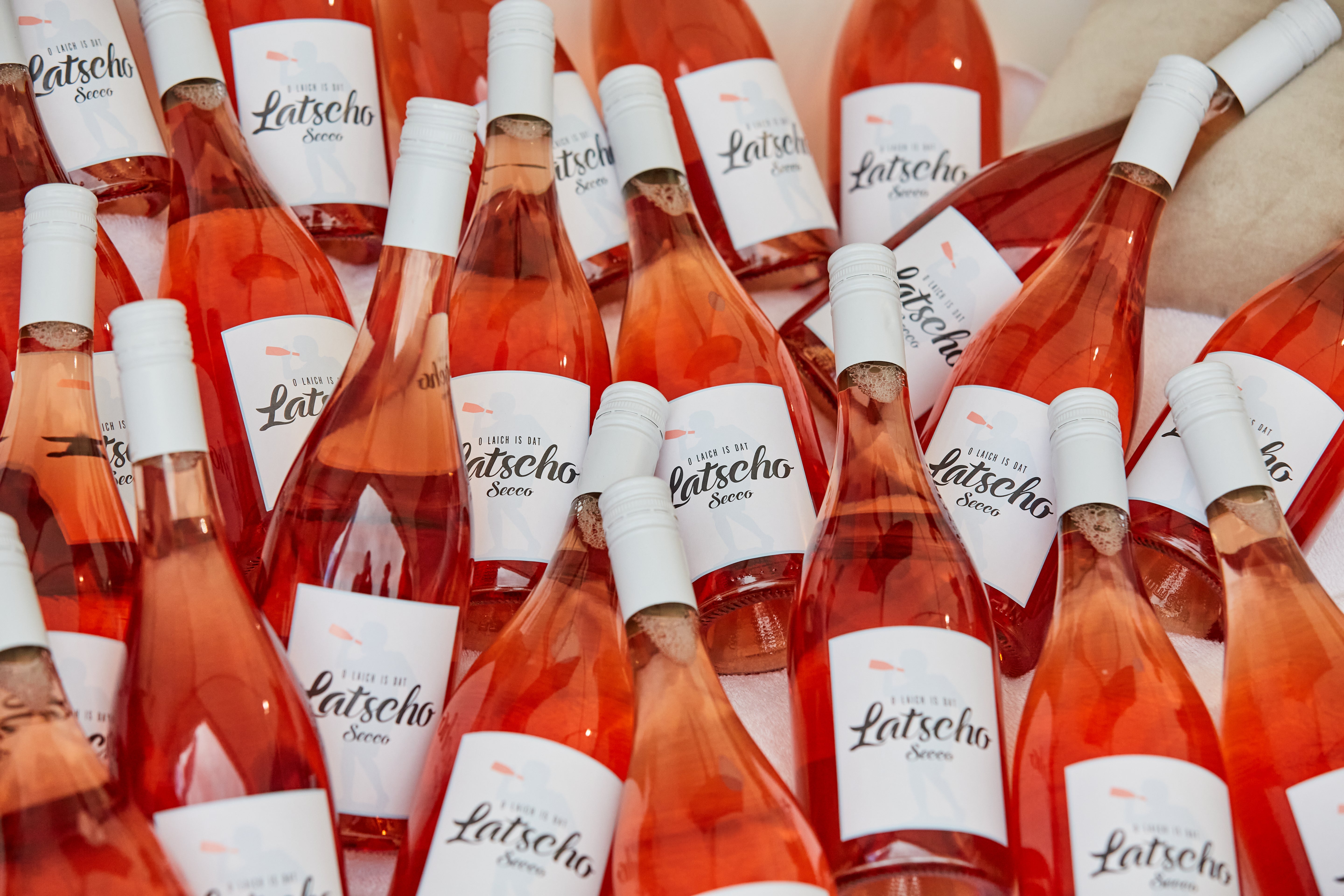 Mehrere Flaschen eines Rosé-Getränks mit dem Namen 'Latscho Secco' liegen eng beieinander. Die Etiketten zeigen eine stilisierte Silhouette und den Schriftzug 'O laich is dat latscho secco'.