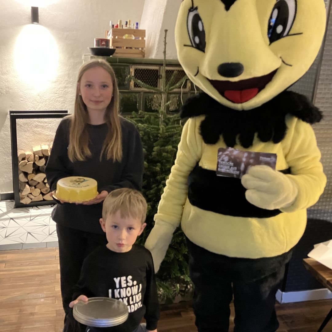Summi, eine Biene und das Maskottchen der Rhein-Zeitung, zu Besuch bei Leni und Max. Im Wohnzimmer der beiden mit den Geschenken: Kuchen, Kinobox und einem Tierpark Gutschein.