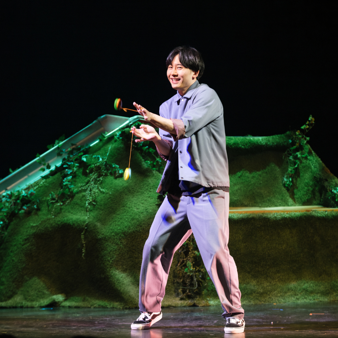 Eine Person zeigt eine Jojo‑Jonglage auf einer Bühne, vor einer mit Moos und Pflanzen gestalteten Kulisse.