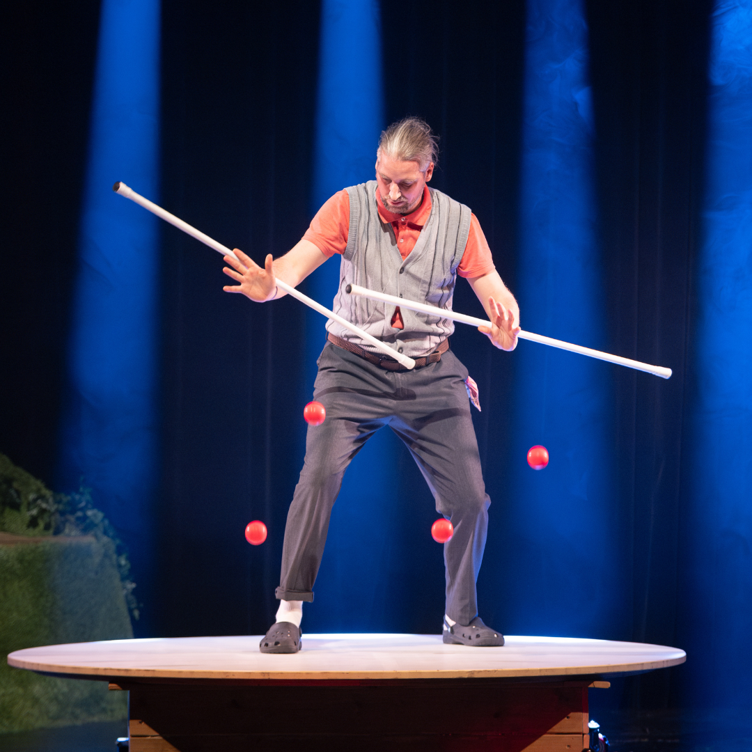 Eine Person jongliert mehrere rote Bälle und balanciert gleichzeitig zwei lange Stäbe auf einer runden Plattform in einer Bühnenbeleuchtung.