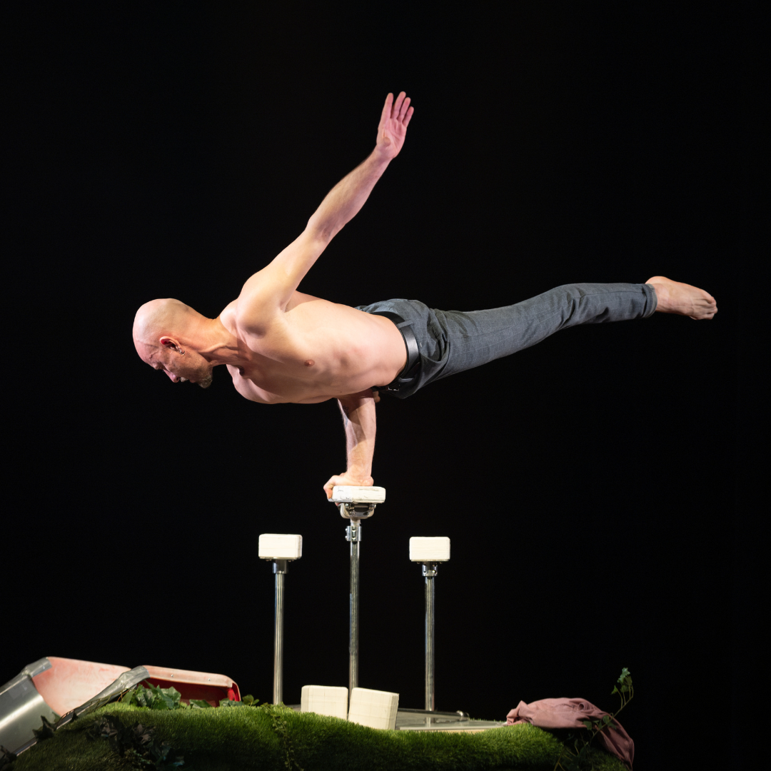 Eine Person balanciert waagerecht in einer einarmigen Handstandposition auf erhöhten Handstandblöcken, vor einem dunklen Bühnenhintergrund.