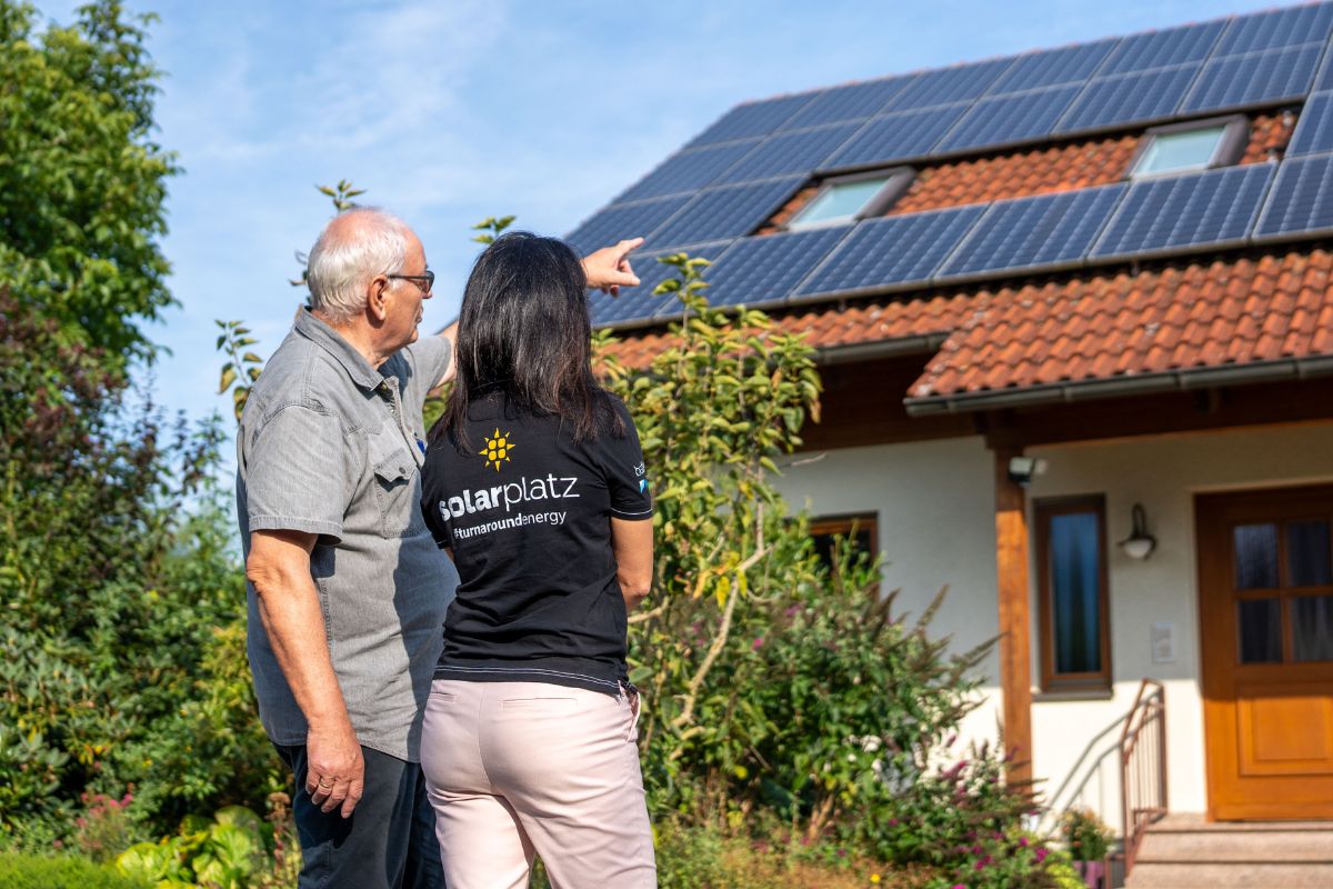 Eine Person zeigt einer anderen Person eine auf dem Hausdach installierte Solaranlage, während beide vor einem Einfamilienhaus mit Garten stehen.