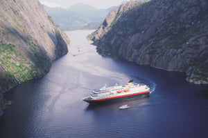 Ein Hurtigruten-Schiff fährt durch eine schmale norwegische Fjordpassage, umgeben von steilen Felswänden, die mit grüner Vegetation bedeckt sind.