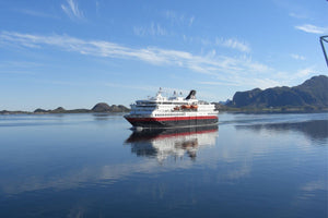 Ein Hurtigruten-Schiff fährt durch einen spiegelglatten Fjord in Norwegen. Das Schiff ist weiß und rot gestrichen und spiegelt sich deutlich im ruhigen Wasser.