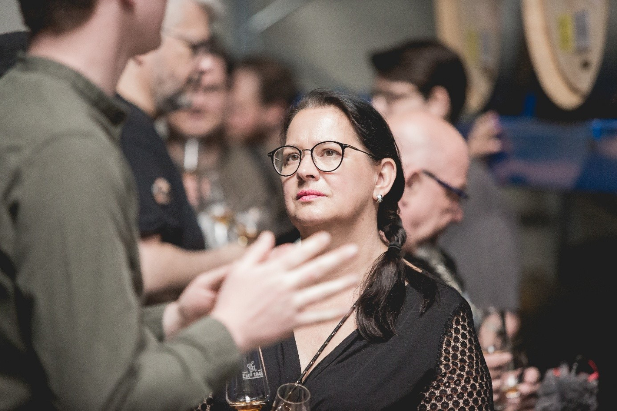 Mehrere Personen unterhalten sich bei einem Whiskey-Tasting mit Gläsern in der Hand; im Hintergrund sind Holzfässer zu sehen.