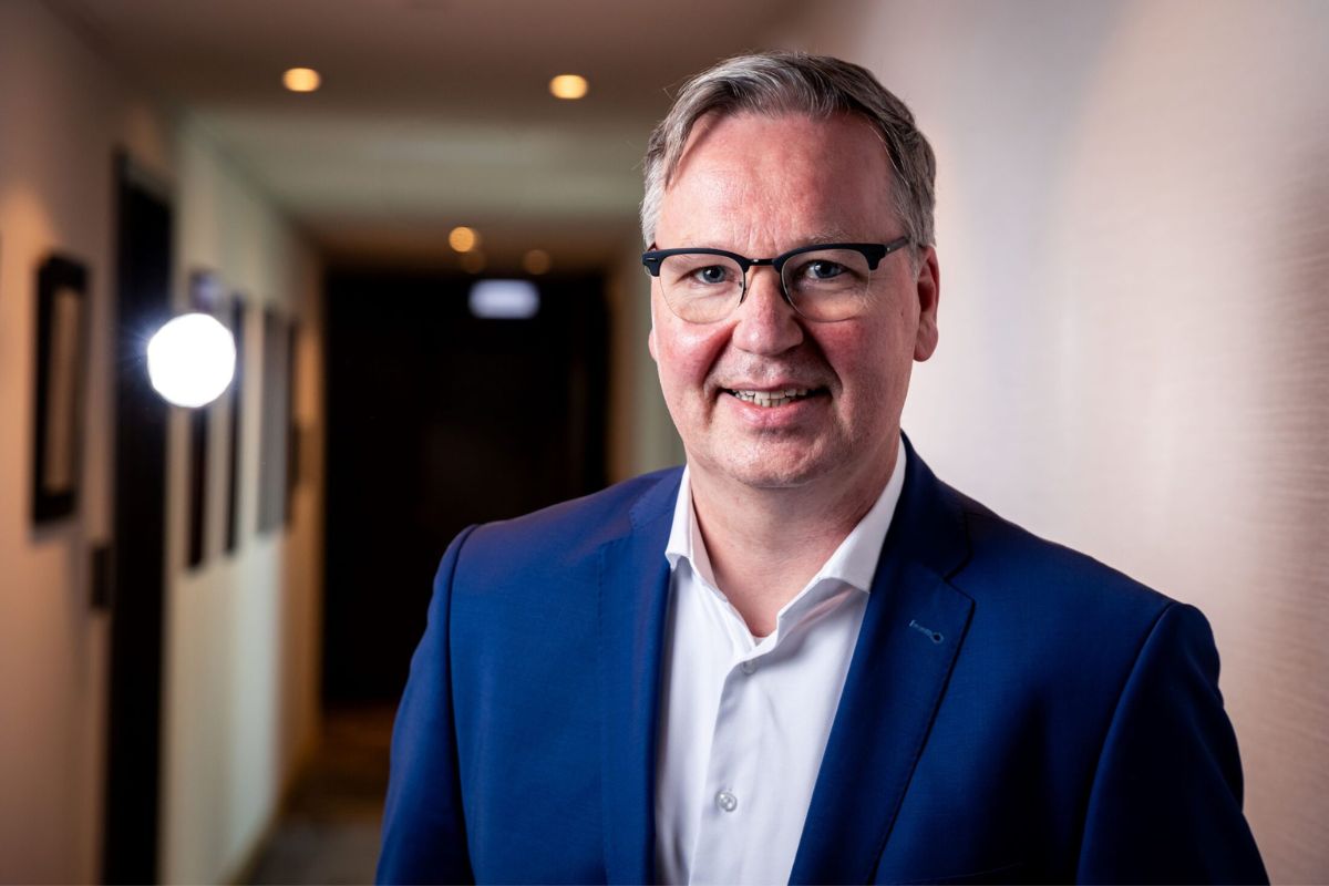 Mann in blauem Anzug und Brille lächelt in die Kamera, stehend in einem beleuchteten Flur.