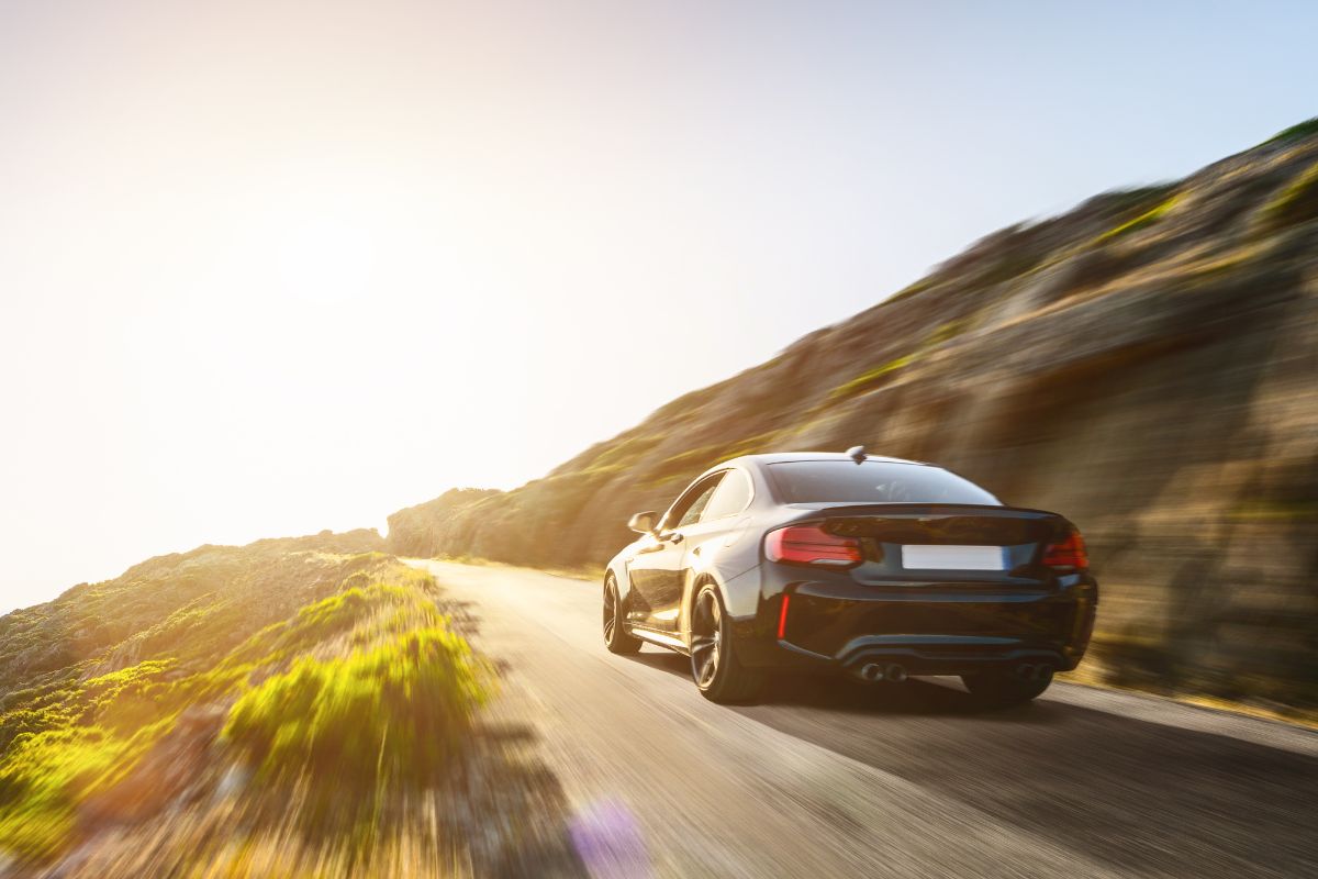 Schwarzes Auto fährt dynamisch auf einer sonnigen Küstenstraße mit Felsen und grünen Landschaften.