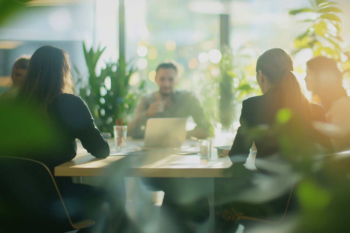 Gruppe von Personen in einem modernen Büro bei einer Besprechung, umgeben von großen Fenstern und grünen Pflanzen
