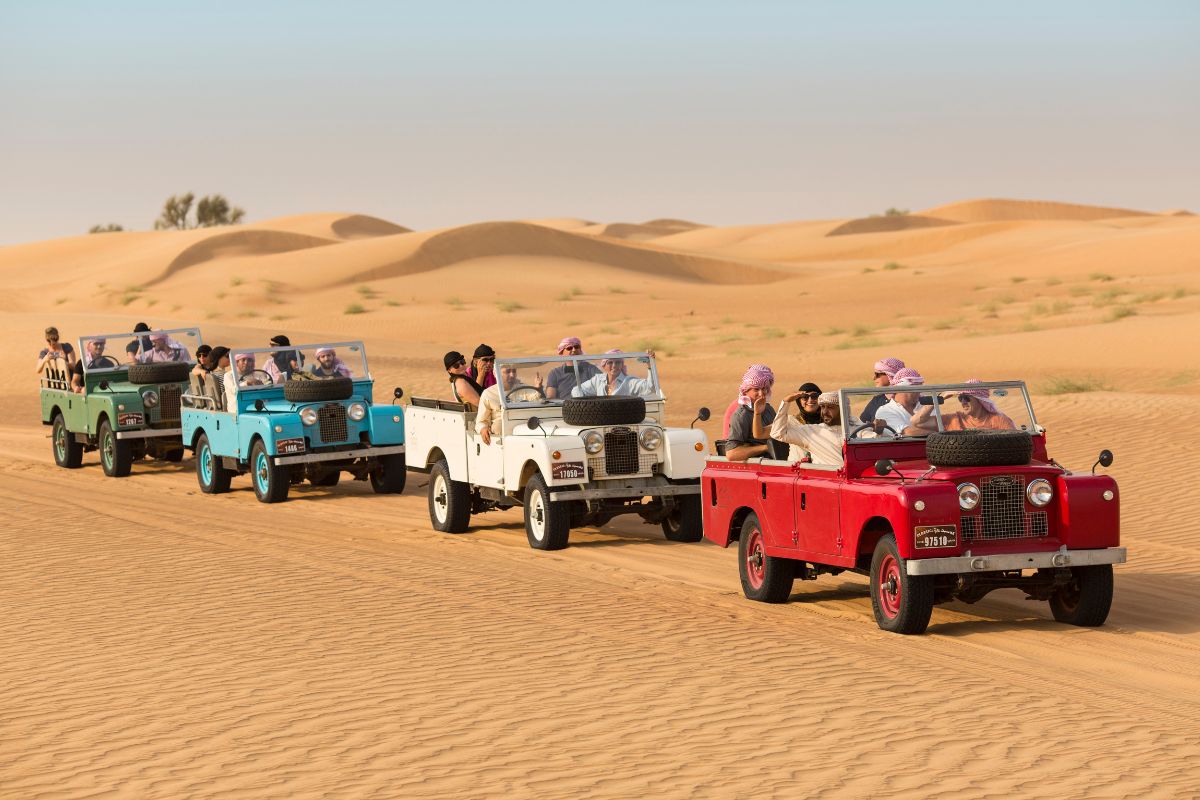 Eine Gruppe von Touristen fährt in mehreren Oldtimer-Geländewagen durch die goldenen Sanddünen der Wüste in Dubai. Die Fahrer tragen traditionelle Kopfbedeckungen und genießen die atemberaubende Landschaft der Wüste.