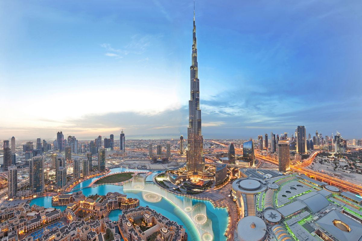 Panoramablick auf den Burj Khalifa, das höchste Gebäude der Welt, inmitten der modernen Skyline von Dubai. Die Szene zeigt auch die umliegenden Gebäude, die beleuchteten Straßen und die Wasserspiele der Dubai Fountain bei Dämmerung.