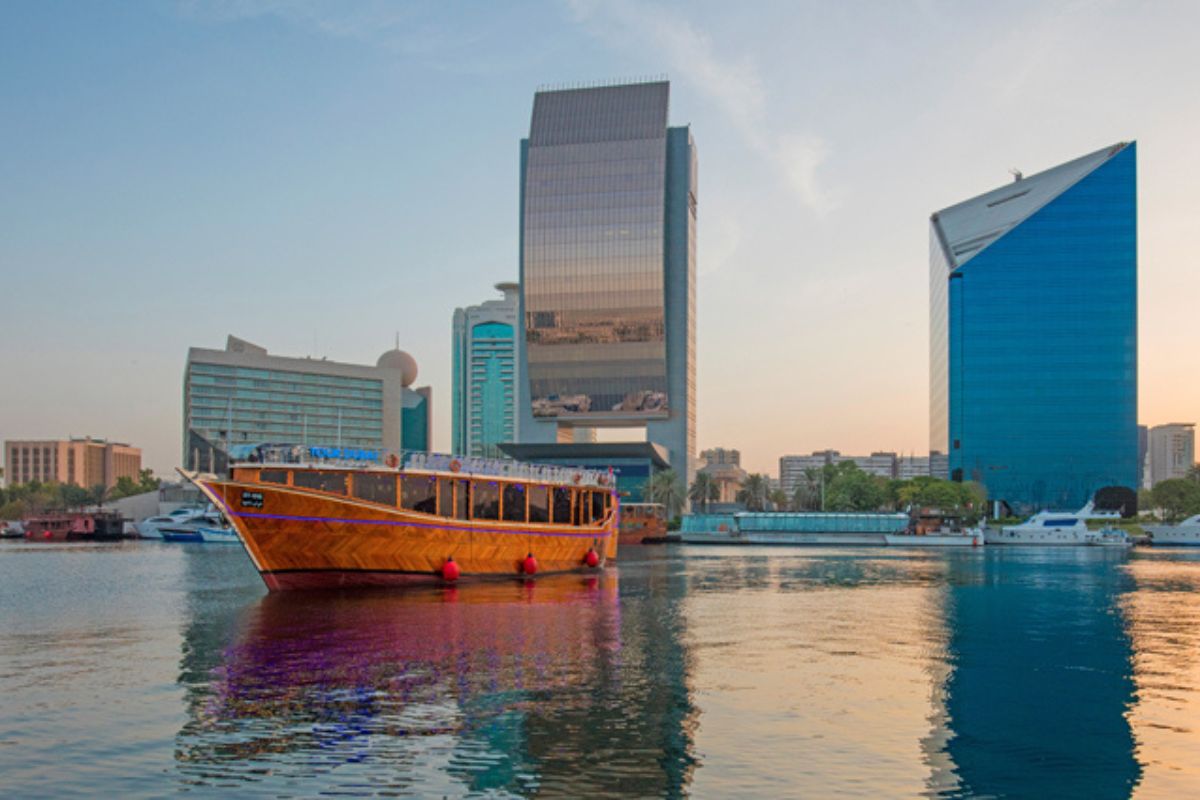 Eine traditionelle arabische Dhow, ein Holzschiff, segelt ruhig auf dem Wasser des Dubai Creek, umgeben von modernen Hochhäusern. Die Skyline mit ihren glänzenden Wolkenkratzern spiegelt sich im Wasser und sorgt für eine beeindruckende Mischung aus Tradition und Moderne.