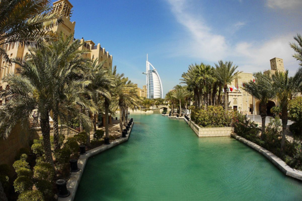 ine malerische Aussicht auf das ikonische Burj Al Arab in Dubai, umgeben von grünen Palmen. Im Vordergrund fließt ein ruhiger türkisfarbener Wasserkanal zwischen den sandfarbenen Gebäuden, der einen Kontrast zur modernen Architektur des Burj Al Arab bildet.