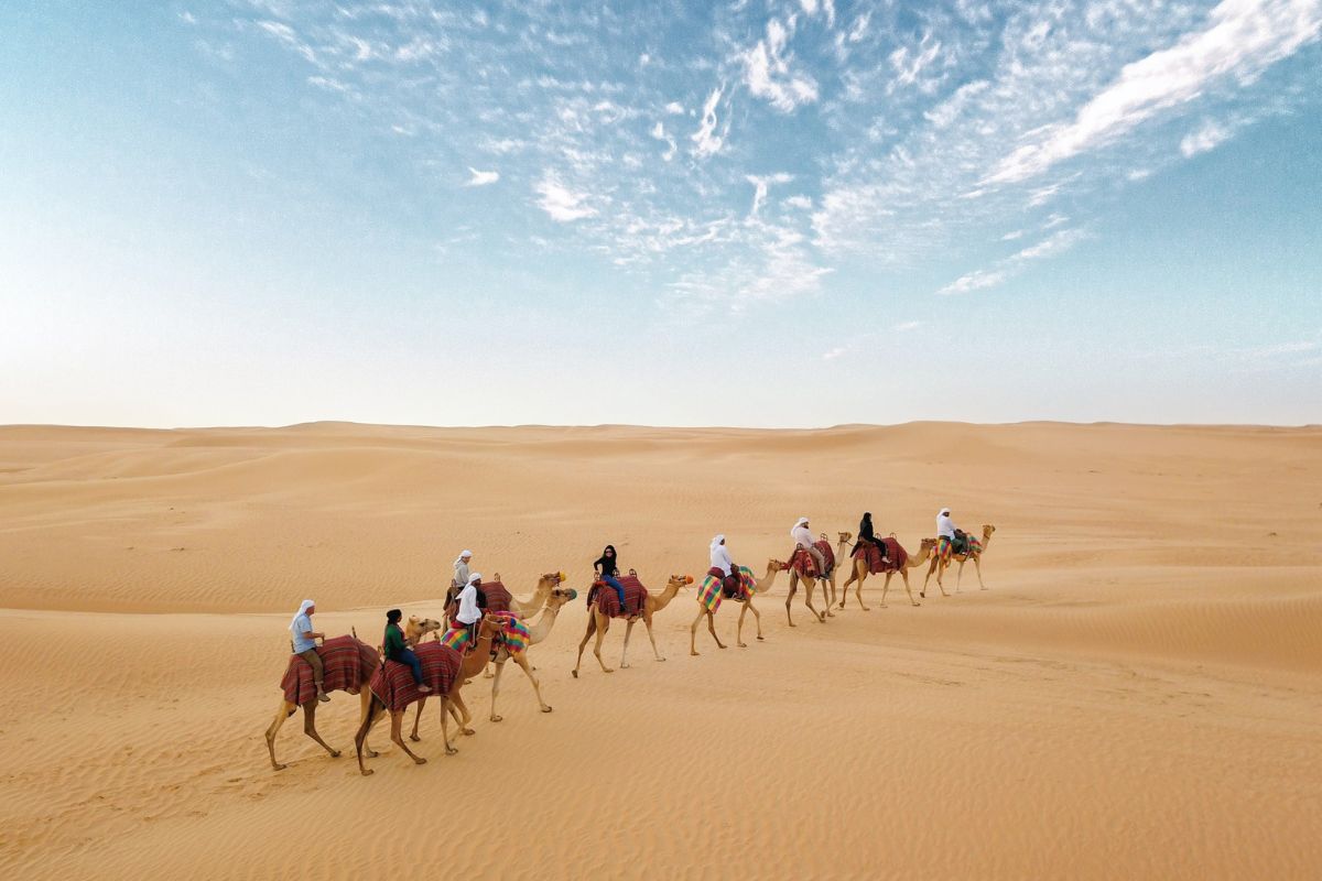 Eine Gruppe von Menschen reitet auf Kamelen durch die weitläufige, sandige Wüstenlandschaft unter einem klaren, leicht bewölkten Himmel. Die Kamele sind mit bunten Decken geschmückt, und die Szene vermittelt ein Gefühl von Ruhe und Abenteuer in der Wüste.