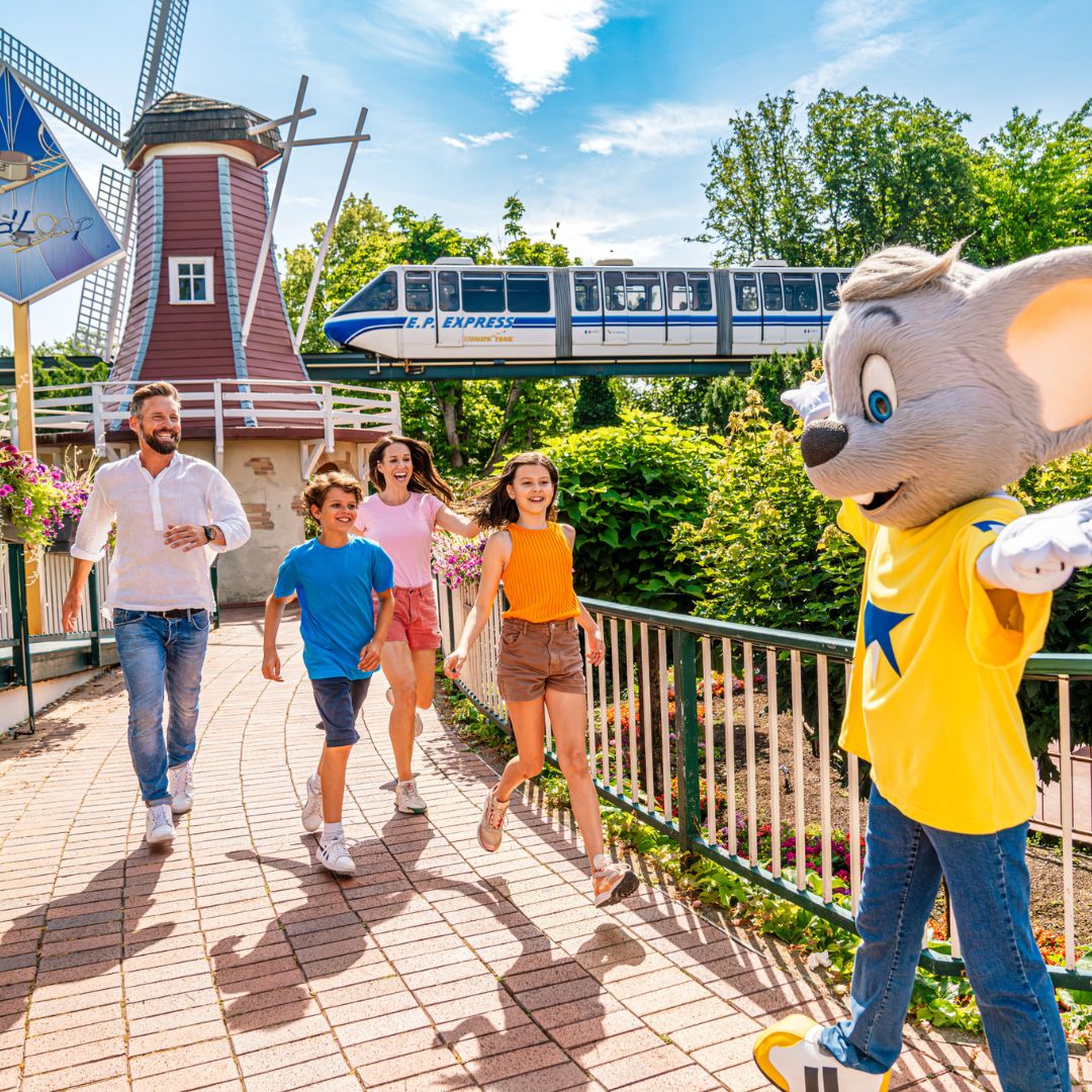 Familie im Europa-Park läuft auf Maskottchen zu, im Hintergrund der E.P. Express und eine Windmühle in grüner Parklandschaft.