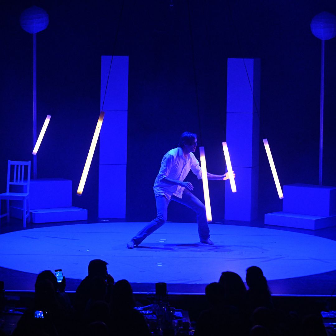 Ein Künstler jongliert mit leuchtenden Stäben auf einer blau beleuchteten Bühne vor Zuschauern.