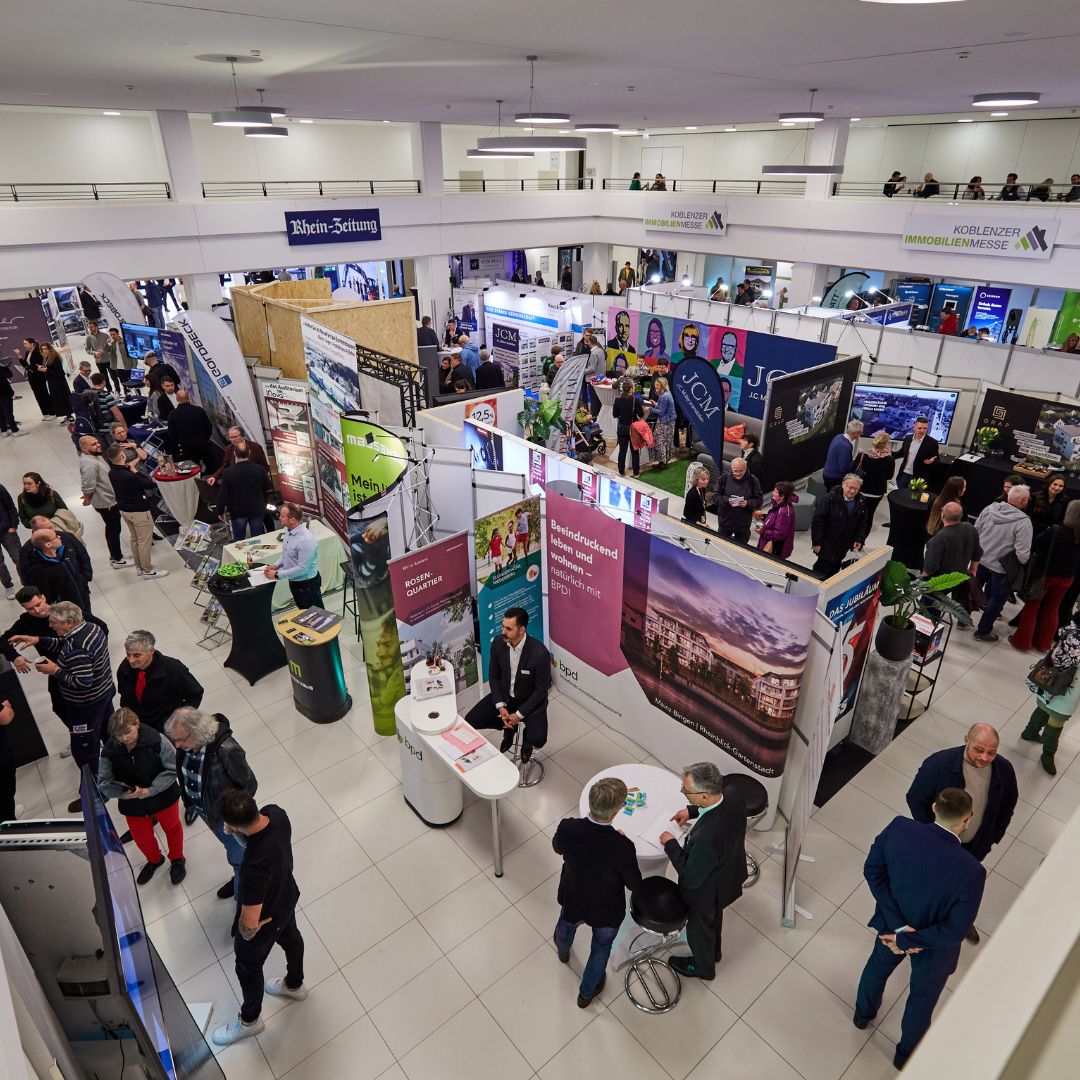 Das Bild zeigt eine belebte Immobilienmesse in Koblenz mit zahlreichen Ständen und vielen Besuchern. Verschiedene Aussteller präsentieren ihre Angebote, und es sind zahlreiche Informationsbanner zu sehen. 