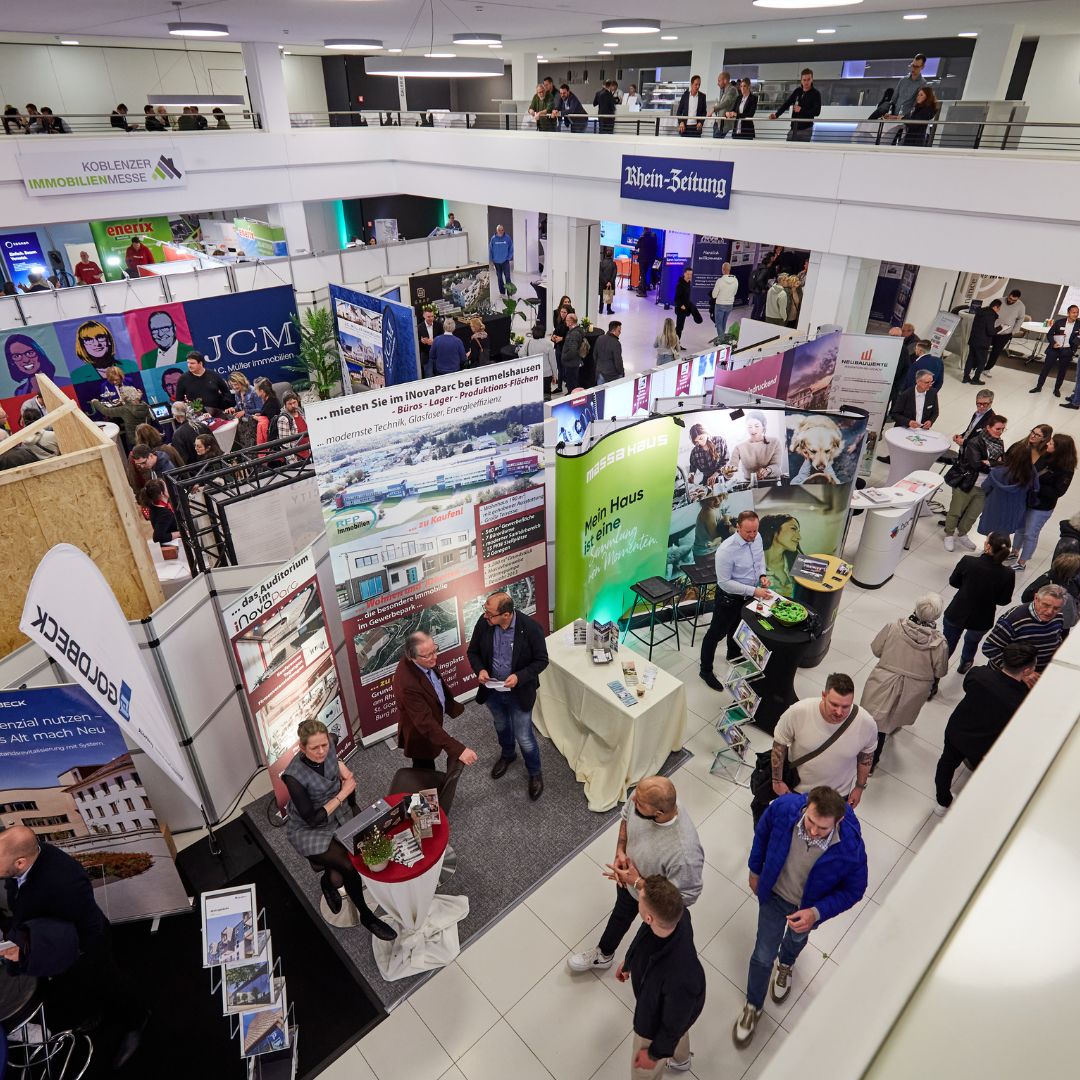 Das Bild zeigt eine belebte Immobilienmesse in Koblenz mit mehreren Ständen und vielen Besuchern, die sich informieren und unterhalten. Ein großes Schild der "Rhein-Zeitung" ist sichtbar, und die Atmosphäre wirkt geschäftig und lebhaft.