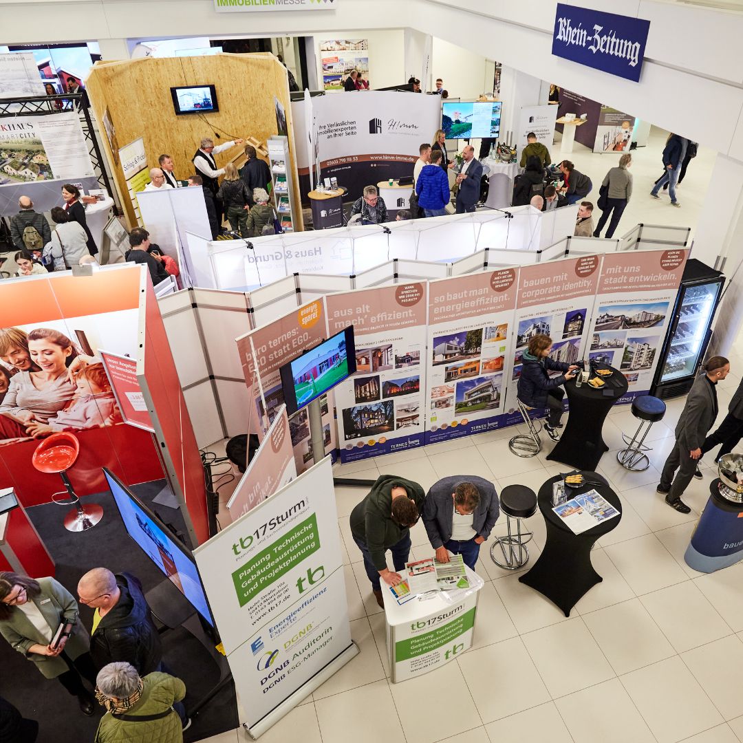 Das Bild zeigt eine belebte Immobilienmesse in Koblenz mit mehreren Ständen, an denen sich Besucher informieren. Verschiedene Informationsstände und Werbebanner sind zu sehen. Ein großes Schild der "Rhein-Zeitung" hängt über dem Bereich. Menschen unterhalten sich, blättern durch Broschüren und informieren sich über Immobilienangebote. Die Atmosphäre ist geschäftig und lebhaft.