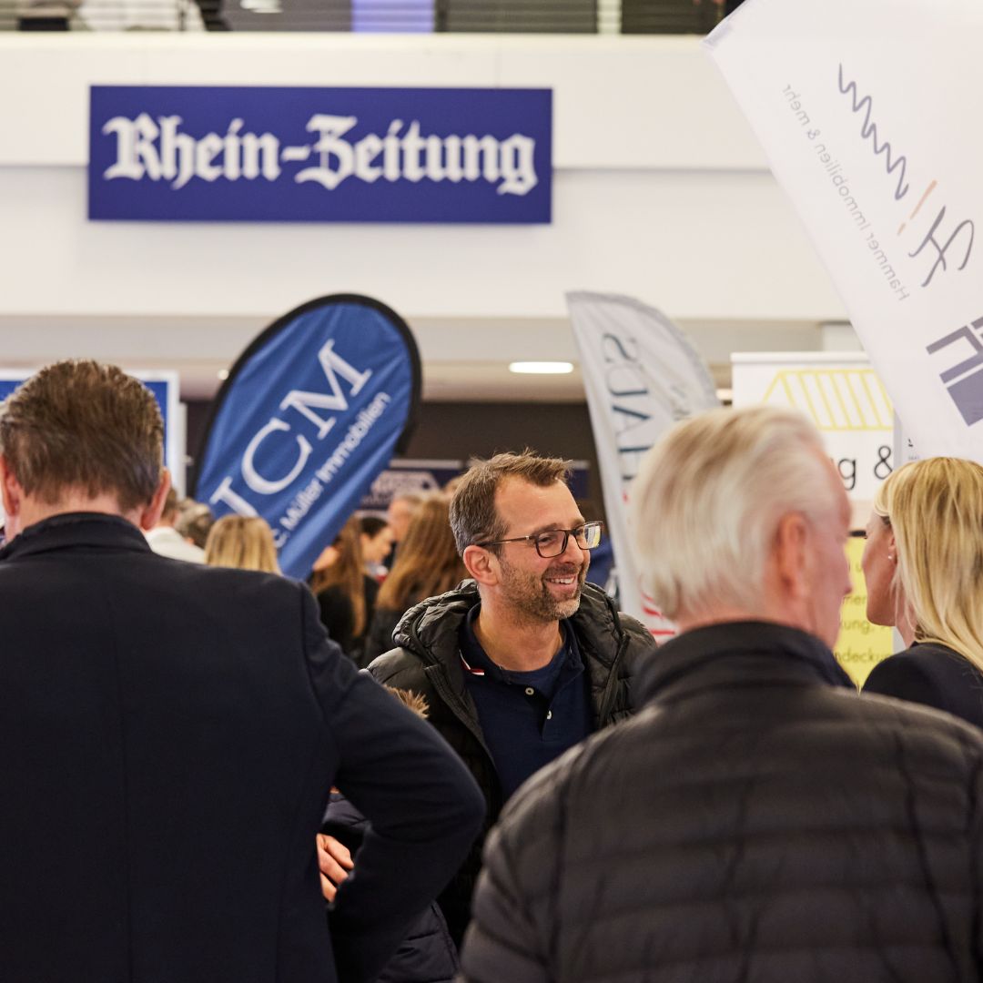Das Bild zeigt eine Szene von einer Immobilienmesse in Koblenz. Im Vordergrund unterhalten sich mehrere Personen lebhaft. Ein Mann mit Brille lächelt und spricht mit einer Frau. Im Hintergrund sind weitere Besucher sowie verschiedene Firmenbanner und ein großes Schild der "Rhein-Zeitung" zu sehen. Die Atmosphäre wirkt freundlich und geschäftig.