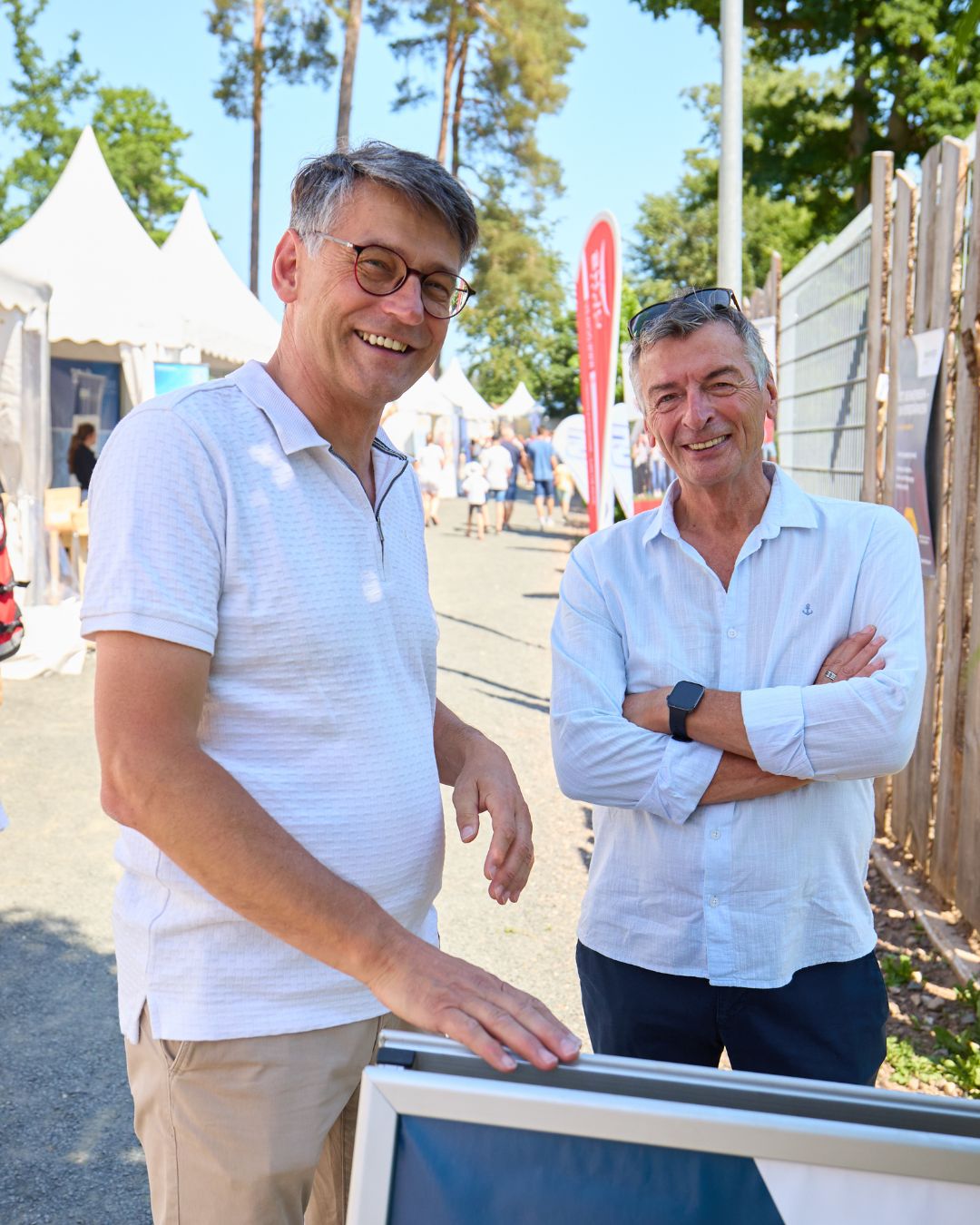 Zwei Personen stehen an einem Kundenstopper bei der Jobmesse „Jobs im Park“ und blicken in die Kamera. Im Hintergrund befinden sich weitere Messestände und Besucher.