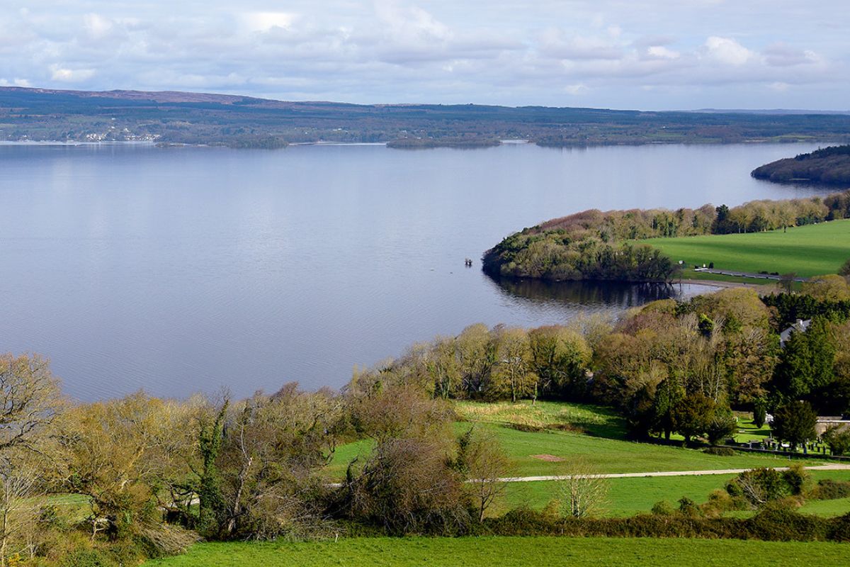 Panoramablick auf einen ruhigen, großen See umgeben von einer vielfältigen Landschaft, die von grünen Wiesen, dichten Wäldern und fernen Hügeln geprägt ist.