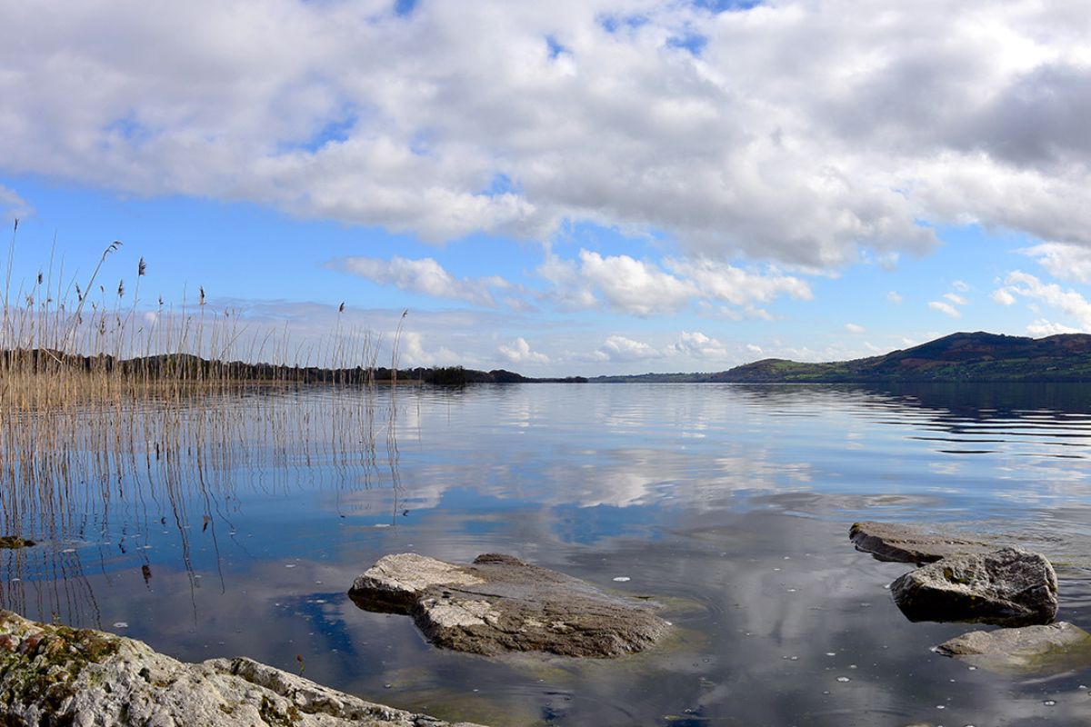 Idyllische Seeansicht mit klarem Wasser, das den Himmel reflektiert, Schilf am Ufer und großen Steinen im Vordergrund. Im Hintergrund hügelige Landschaft unter einem leicht bewölkten Himmel.