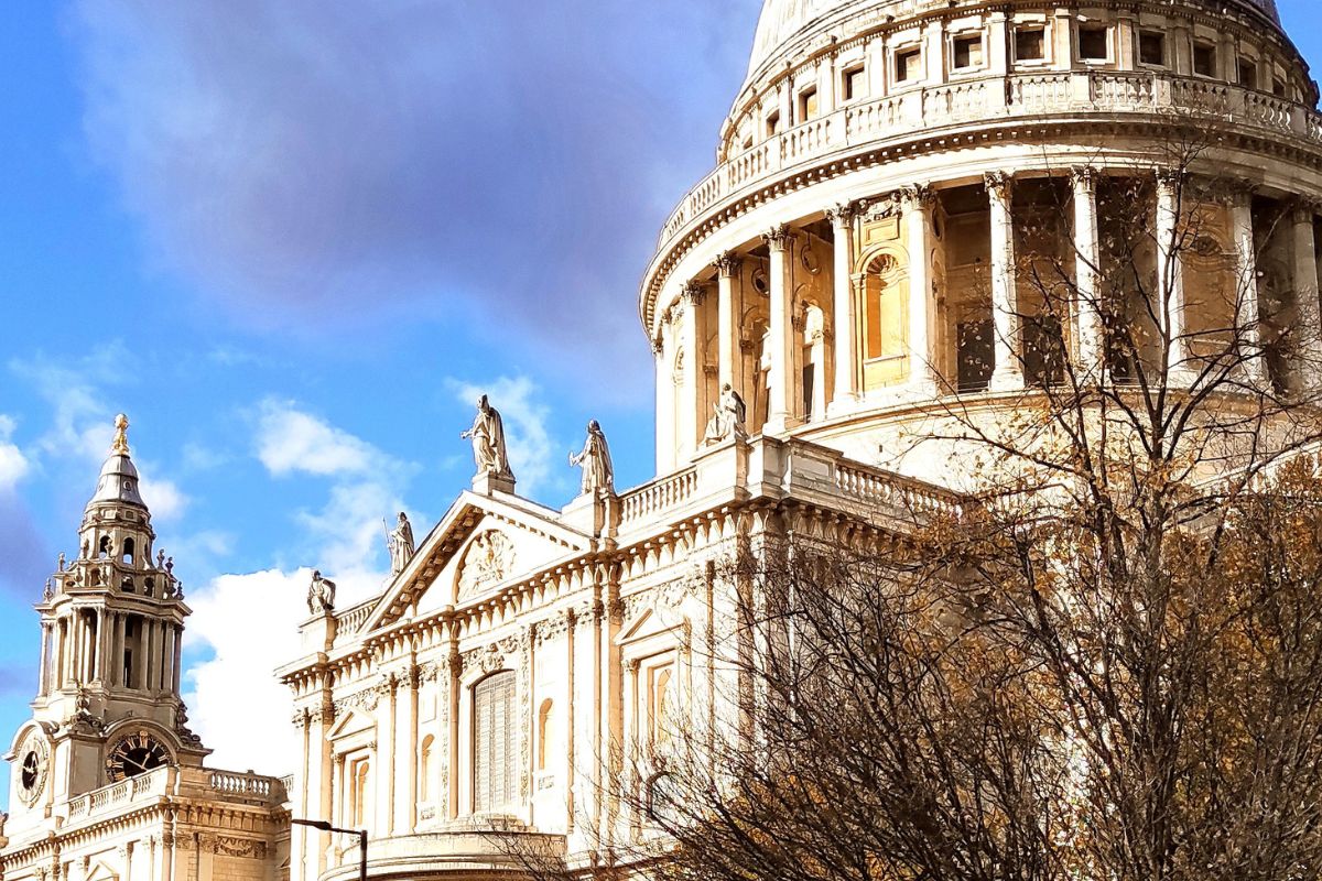 St. Paul’s Cathedral in London unter blauem Himmel mit leichten Wolken. Die markante Kuppel steht im Vordergrund, begleitet von Bäumen und einem roten Doppeldeckerbus auf der Straße.