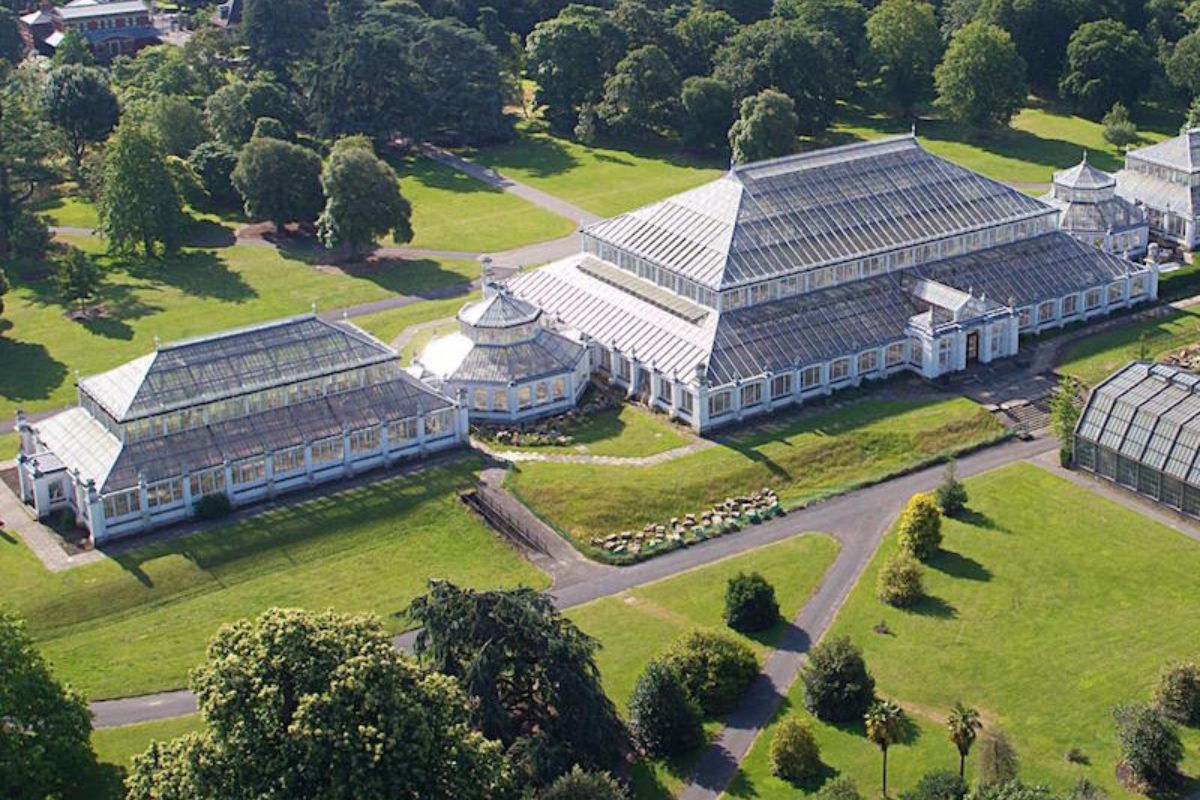 Eine beeindruckende Luftansicht des Temperate House in den Kew Gardens, London. Das riesige viktorianische Gewächshaus mit seinen gläsernen Dächern und umgeben von grünen Rasenflächen und Bäumen.