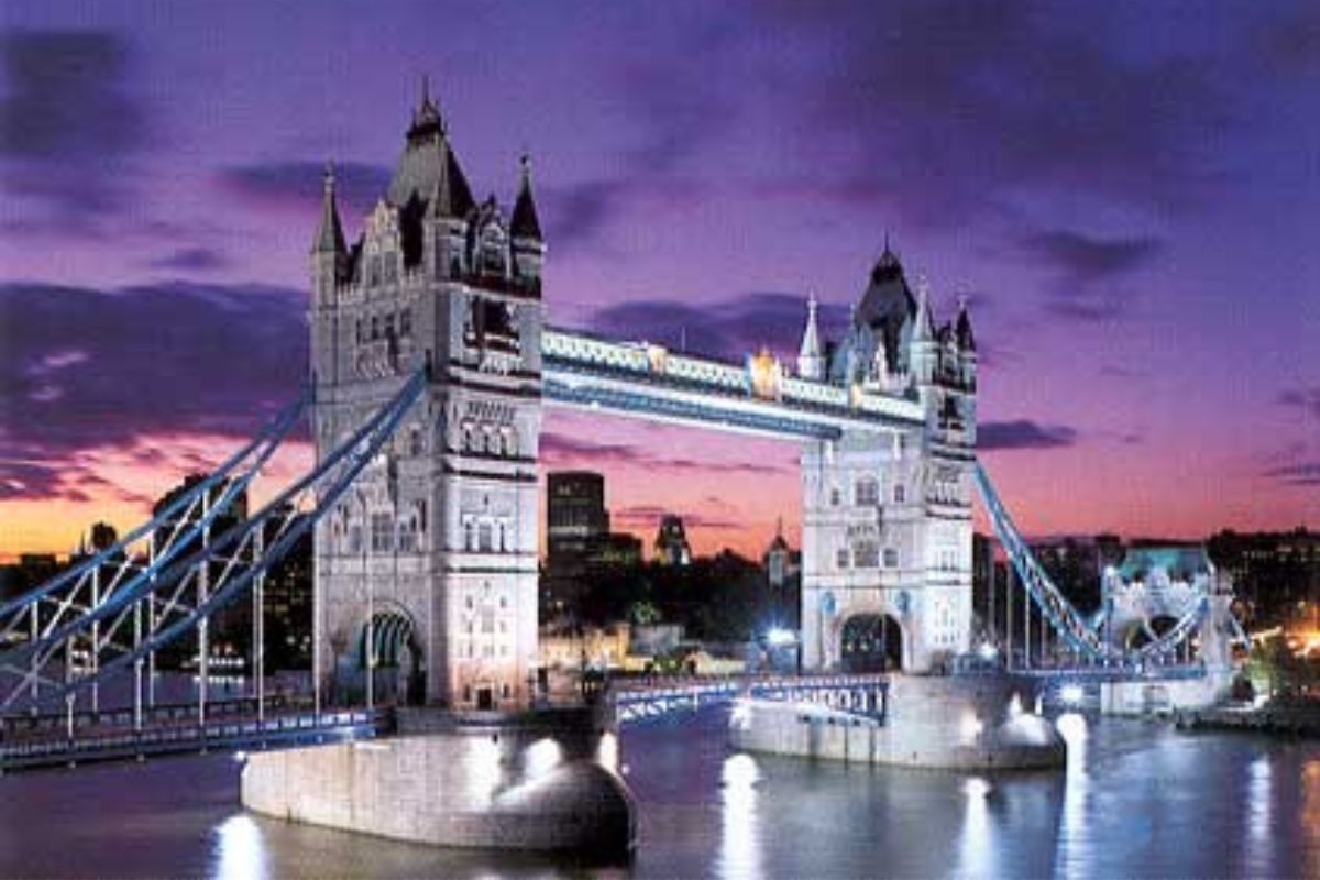 Ein abendlicher Blick auf die beleuchtete Tower Bridge, die sich über die Themse spannt. Der Himmel ist in tiefen Lilatönen gefärbt, während die Brücke hell erleuchtet ist und sich ihre Reflexion im ruhigen Wasser darunter widerspiegelt. Im Hintergrund sind Teile der Londoner Skyline bei Dämmerung zu erkennen.