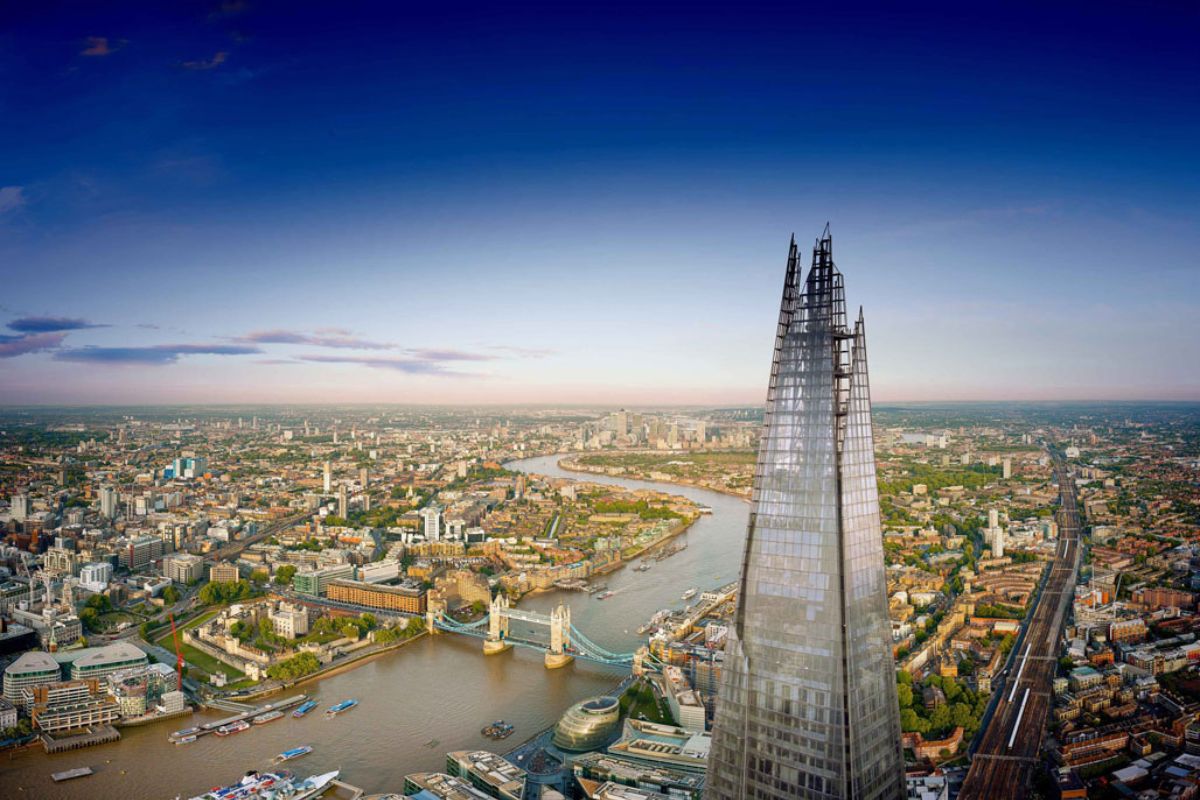 Ein atemberaubender Blick auf den Shard, eines der höchsten Gebäude in London, der sich über die Stadt erhebt. Im Hintergrund fließt die Themse, und die Tower Bridge ist zu sehen, zusammen mit den umliegenden Gebäuden und Straßen. Die Skyline erstreckt sich bis zum Horizont unter einem klaren blauen Himmel.