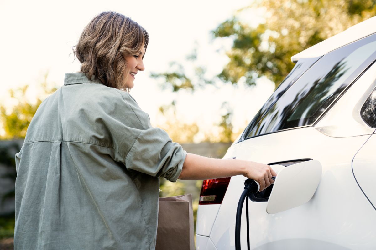 Eine junge Frau lädt ihr Elektroauto auf. Sie steht lächelnd neben dem Auto und hält das Ladekabel in der Hand.