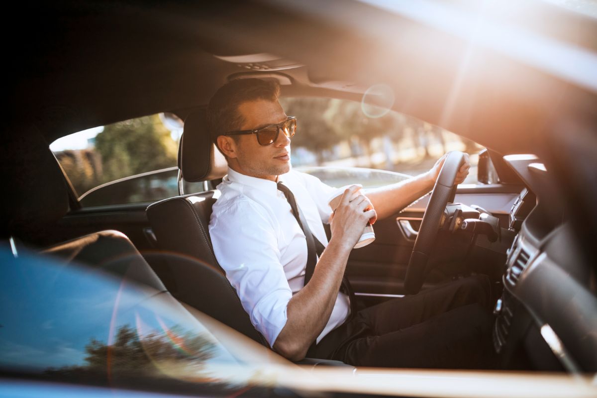 Ein Geschäftsmann sitzt in einem Auto, hält einen Kaffeebecher und trägt eine Sonnenbrille. Er wirkt entspannt und genießt die Fahrt.