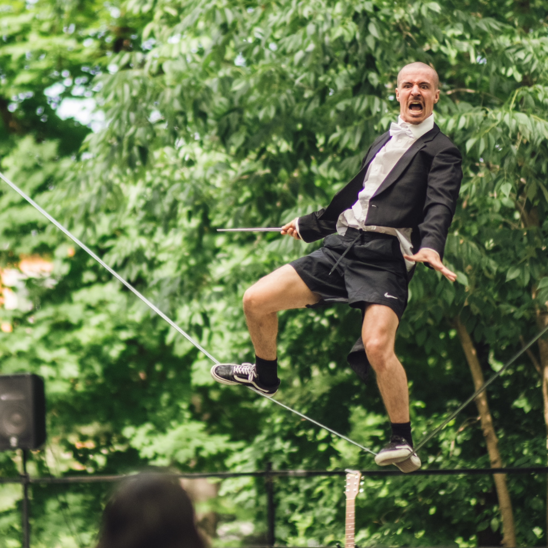 Eine Person balanciert auf einem Drahtseil im Freien und trägt dabei ein formelles Oberteil kombiniert mit kurzen Hosen, während die Umgebung von grünen Bäumen umgeben ist.