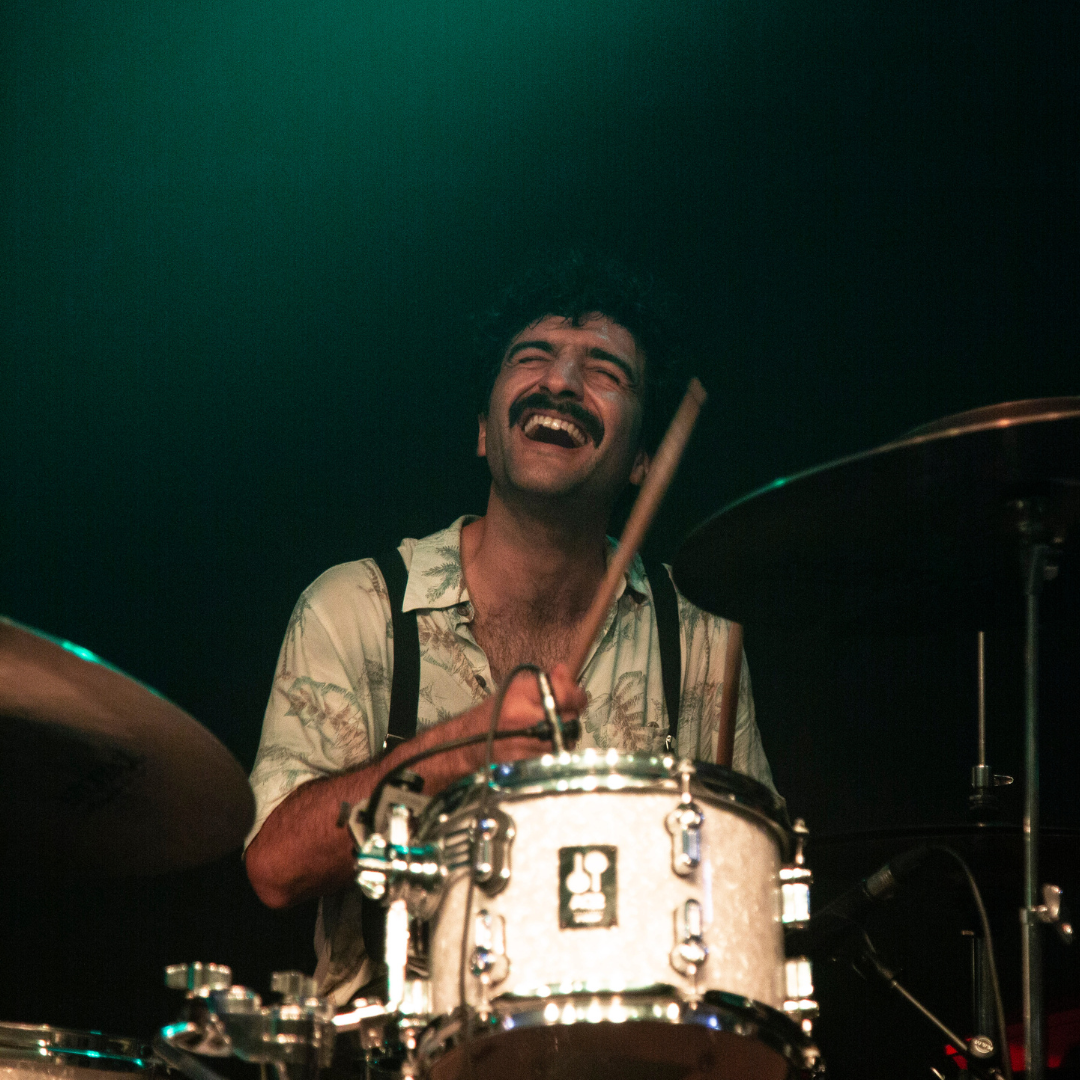 Eine Person spielt Schlagzeug auf einer dunkel beleuchteten Bühne, die Schlagzeugsticks sind in Bewegung und die Umgebung ist in grünliches Licht getaucht