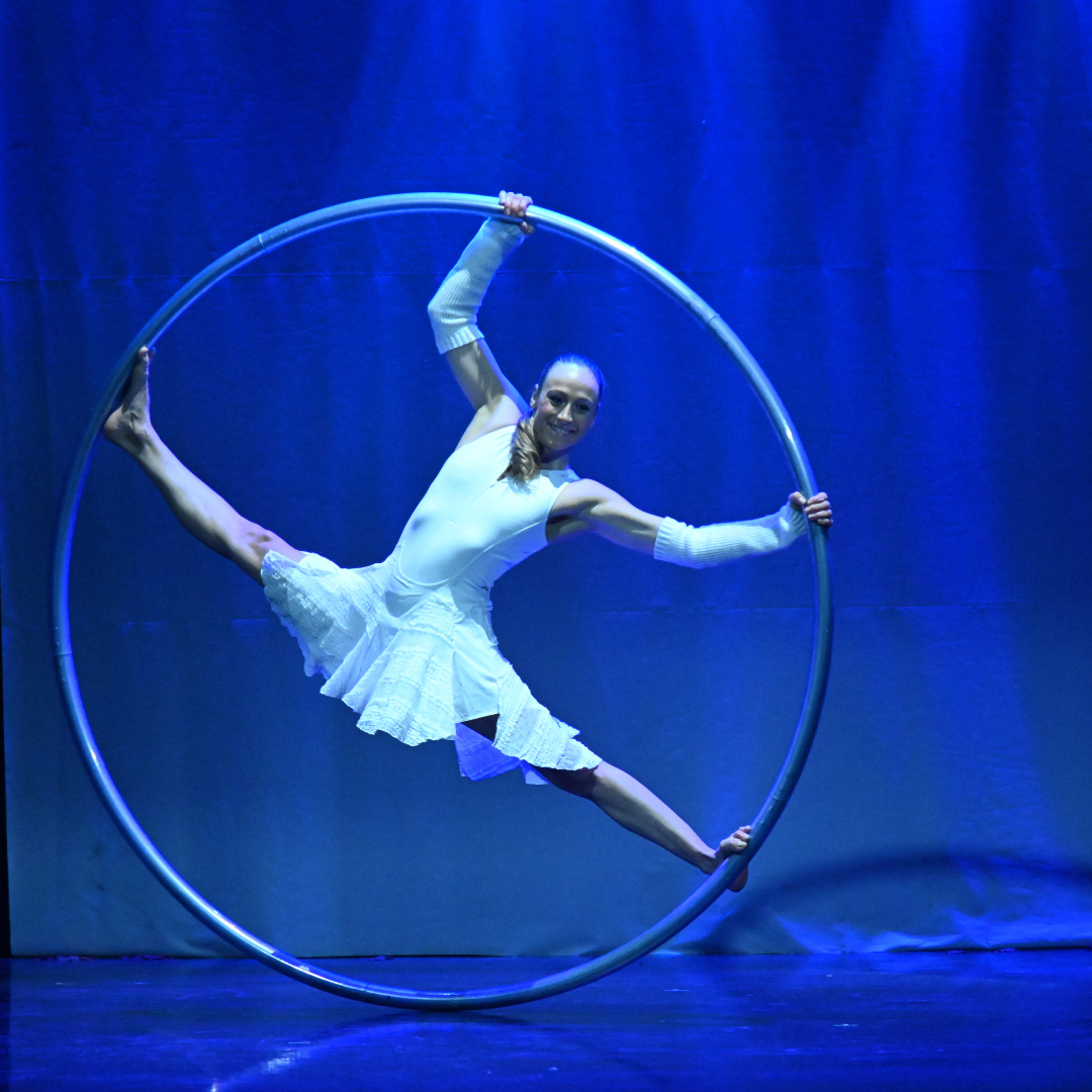 Eine Person führt eine Kunstperformance in einem großen Metallring auf, der auf einer blau beleuchteten Bühne steht.