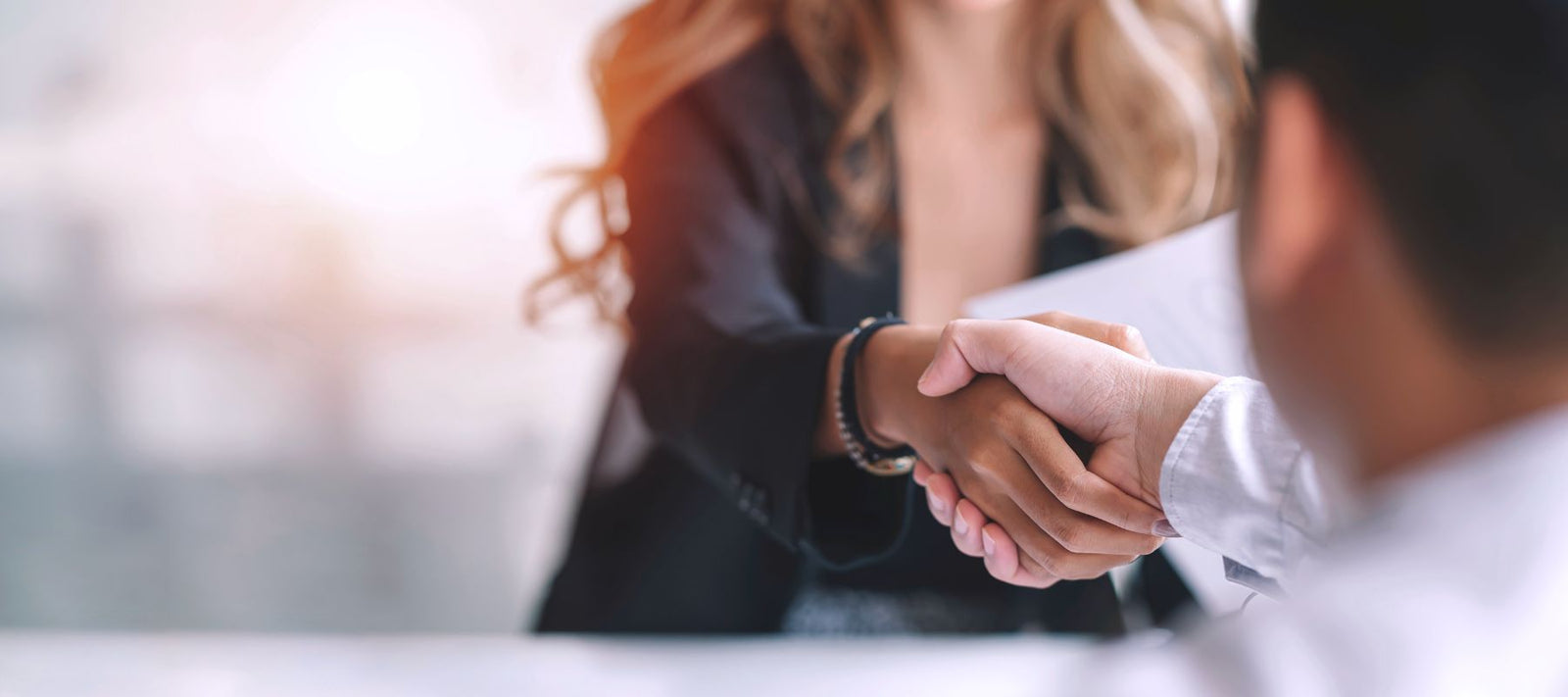 Nahaufnahme eines Händedrucks zwischen zwei Personen in einem formellen Kontext. Eine Frau mit langen Haaren und einem schwarzen Blazer schüttelt die Hand eines Mannes. Der Hintergrund ist verschwommen, was den Fokus auf den Händedruck legt. Die Atmosphäre wirkt professionell und freundlich.